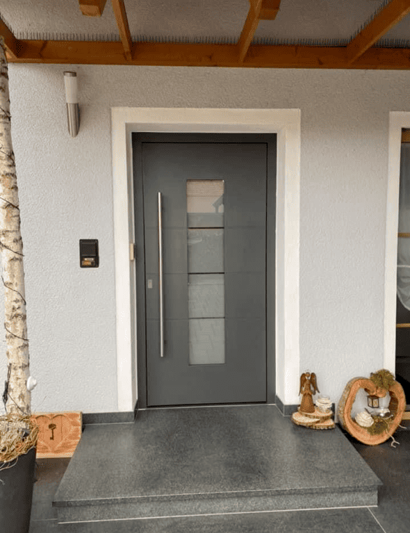 Eine dunkelgraue Haustüre mit 4 gleich großen Glasrechtecken in der Mitte. Zuvor befindet sich eine steinerne Treppenstufe.