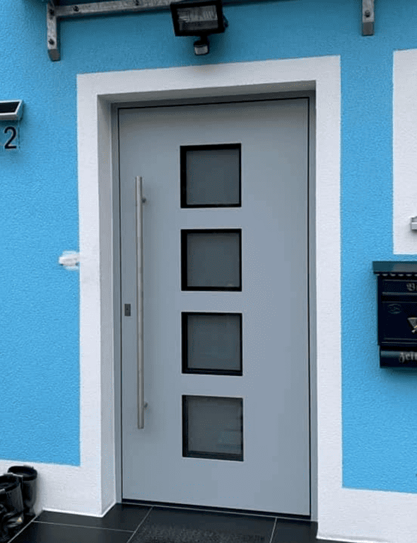 Eine graue Haustüre mit 4 quadratischen Glaselementen, die Hauswand ist hellblau mit weißen Rahmen.