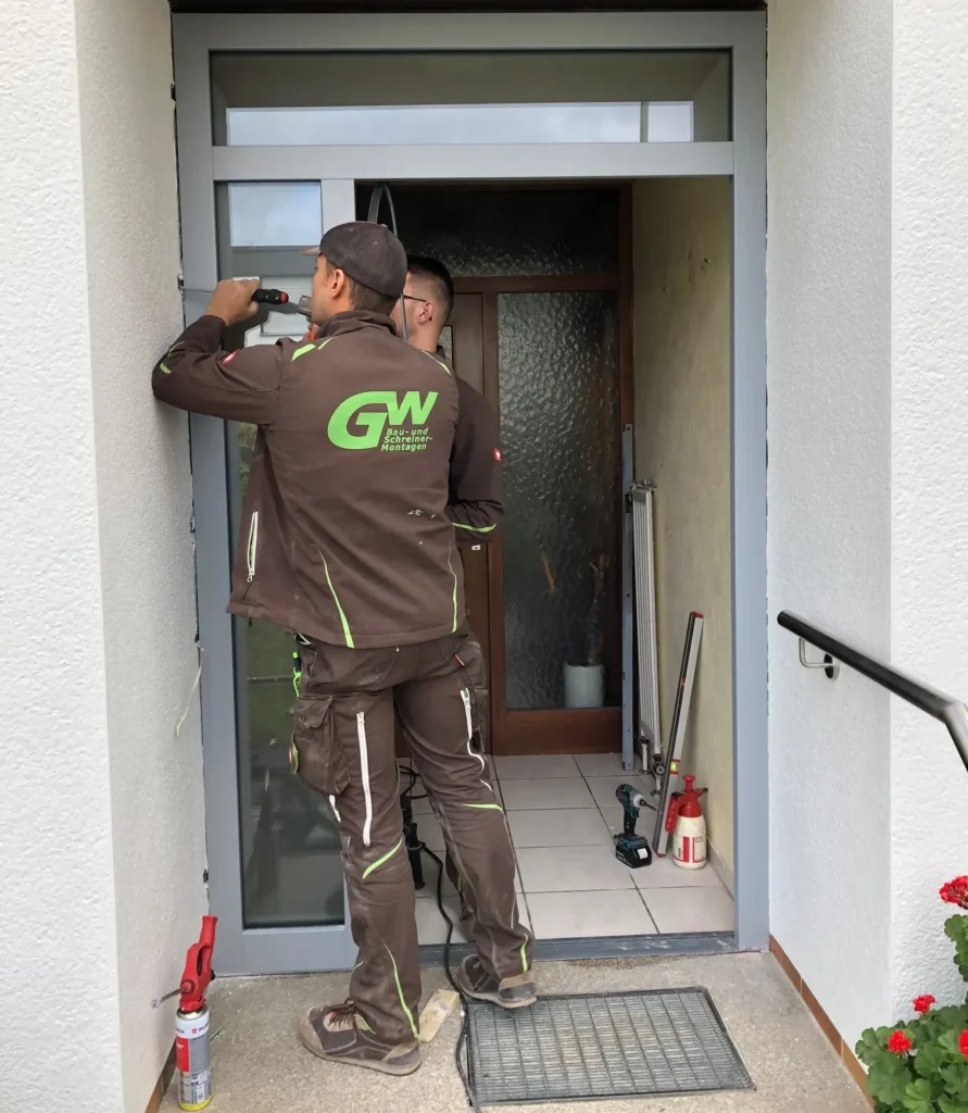 Zwei Männer verfugen gerade die neu gesetzte graue Haustüre mit Glaselementen rechts und oben.