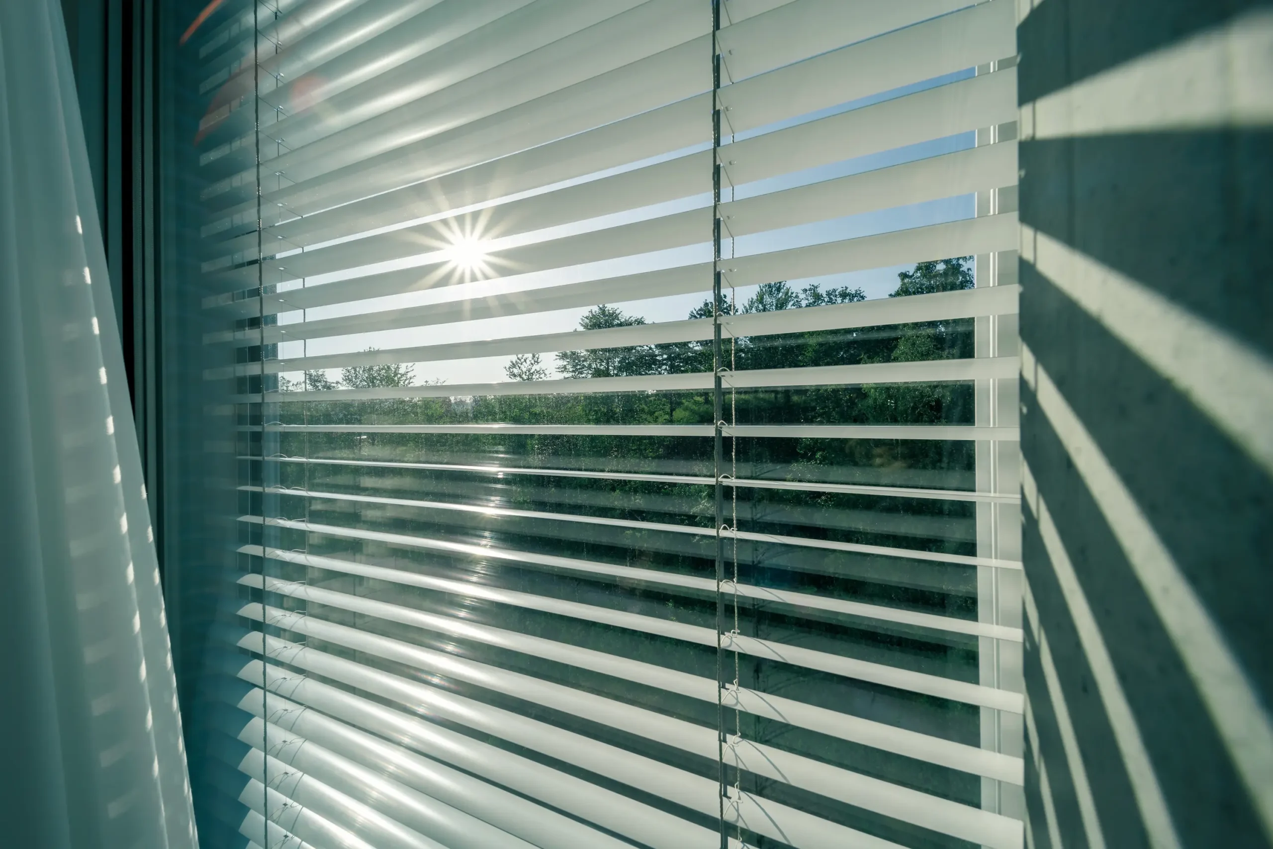 Ein Blick von innen in die Sonne durch eine gekippte, weiße Innenjalousie am Fenster.