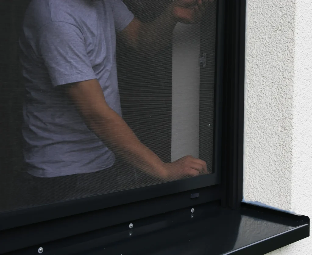 Ein Mann in kurzen Shirt installiert von innen das Insektenschutzgitter am schwarzen Fenster.