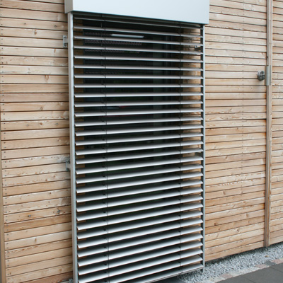 Ein silbernes geschlossenes Raffstore. Die Hausfassade ist aus hellen Holz.