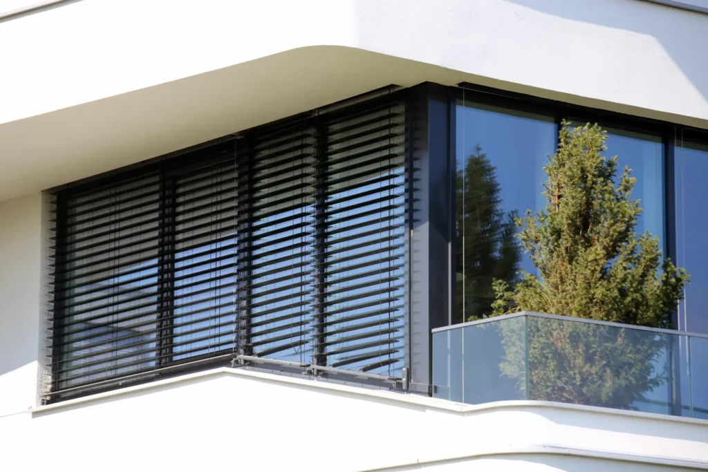 Ein schwarzes Raffstore ist heruntergelassen und gekippt, sodass das Fenster im Hintergrund zu sehen ist.