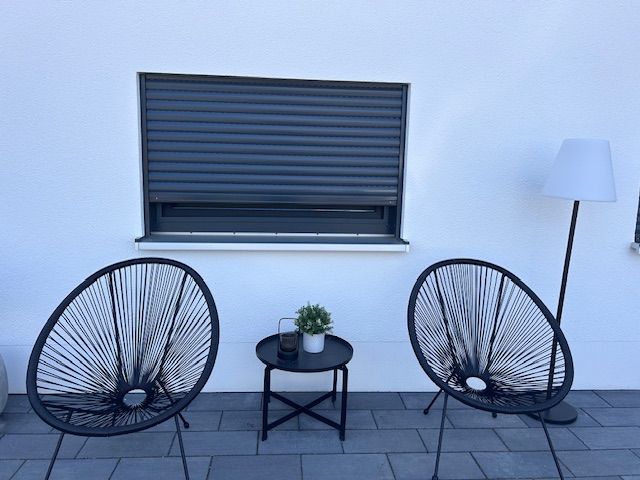 Ein schwarzer Rollo mit zwei schwarzen Stühlen auf einer Terrasse davor.